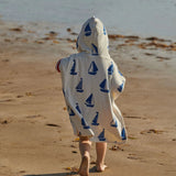 Back view of a person wearing a white hooded towel with blue sailboat patterns on a sandy beach.
