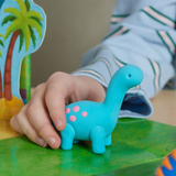 Child's hand holding a blue dinosaur clay toy on a green surface with a colourful card in the background.