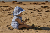 Child sitting on a sandy beach wearing a blue striped hat and matching outfit.