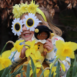 Child wearing sunflower glasses and yellow flower headband, holding the OLLI ELLA Dinky Dinkum Dolls - Billie Bumblebee - Yellow in a bush of daffodils