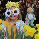 Child wearing sunflower glassses and yellow flower headband holding the OLLI ELLA Dinky Dinkum Dolls - Gideon Grasshopper - Green in the daffodil bush
