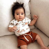 Baby wearing a dinosaur-themed shirt and rust-colored shorts lying on a beige couch.