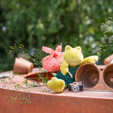 Plush frog toy holding a pink sack and camera on a wooden surface with greenery in the background