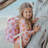 Young girl with a pink backpack featuring strawberry pattern, standing near a wooden log on a beach.