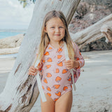 Young girl in a pink swimsuit with orange strawberry pattern standing next to a tree on a beach.