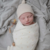 Newborn baby wrapped in a white blanket with yellow suns, wearing a beige knit hat, lying on gray blankets.