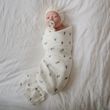 Newborn baby wrapped in a whale print muslin swaddle with a pacifier, lying on a soft surface.