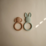 Two silicone teething rings, one brown dog and one green bunny, on a beige background.