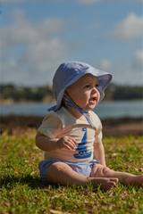 Baby sitting on grass wearing a blue sun hat and shirt with a sailboat design.