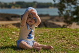 Baby sitting on grass wearing a hat and shirt with a sailboat design.