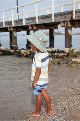 Child wearing a sun hat and striped shirt standing on a beach near a wooden pier.