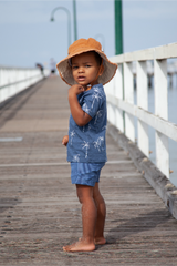 Child wearing a blue outfit with white patterns and a brown hat on a wooden pier.