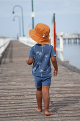 Child wearing a blue shirt and shorts with a sun hat, standing on a wooden pier.