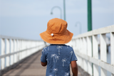 Child wearing an orange hat and blue shirt with palm tree pattern on a wooden pier.