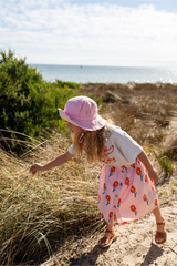 Child in a floral dress and pink hat standing on a beach with ocean view