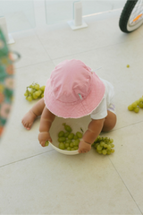 Baby in a pink hat playing with green grapes on a light-colored floor.