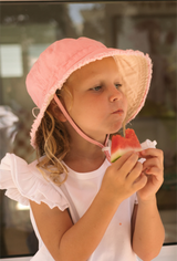 Young girl wearing a pink sun hat eating watermelon outdoors.