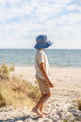 Child wearing a blue sun hat walking on a sandy beach with ocean and sky in the background