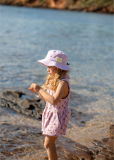 Young girl in a pink dress and lavender hat standing by a body of water.