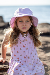 Young girl in a floral dress and lavender hat sitting on a beach.