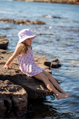 Young girl sitting on rocks by a body of water wearing a pink dress and sun hat.