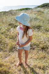 Young girl in a light blue hat and white shirt standing in a grassy field with a coastal background.