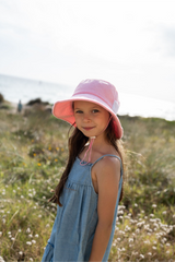 Young girl wearing a pink bucket hat and blue dress standing in a grassy field.