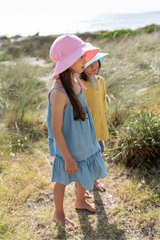 Two children in dresses and hats standing on a grassy path near the beach.