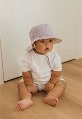Baby sitting on a wooden floor wearing a lavender hat and white shirt.