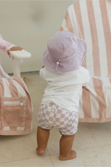 Back view of a Child wearing a lavender hat and checkered shorts standing next to a pink and white striped chair.
