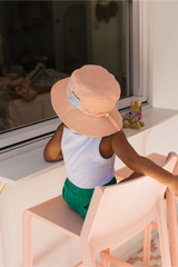 Back view of Child wearing a peach hat sitting on a pink chair by a window.