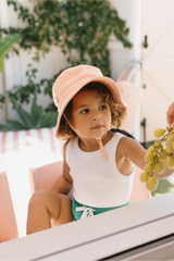 Child wearing a peach sun hat outdoors with greenery in the background