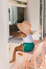 Child sitting on a stool wearing a peach hat and green shorts in an outdoor setting.