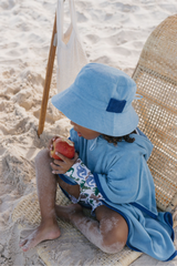 Child in blue towel and hat sitting on a beach with an apple
