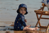 Baby in a blue swimsuit and matching hat sitting in shallow water at the beach.