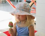 Child wearing a floral hat and blue dress, eating a popsicle on a boat.