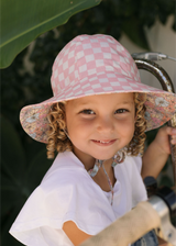 Child wearing a pink checkered sun hat with floral edges, smiling outdoors.