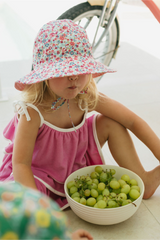 Young girl in a floral hat and pink dress sitting next to a bowl of green grapes.