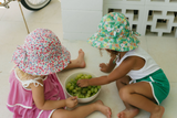 Two children sitting on a white floor, eating grapes from a bowl, wearing floral hats.