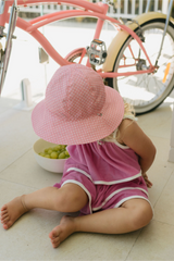 Child wearing a pink sun hat and dress sitting on the floor next to a pink bicycle.