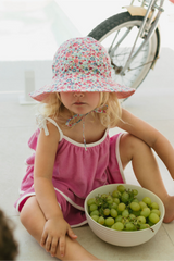 Child in a floral hat and pink dress sitting next to a bowl of green grapes with a bicycle in the background.