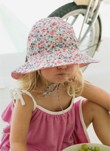 Child wearing a floral sun hat and pink dress, sitting outdoors with a bicycle in the background.