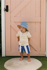 Child standing in front of door wearing a blue hat, cream tee and blue & white striped shorts