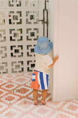 Back view of Child standing next to the outdoor shower wearing a blue hat, cream tee and striped shorts