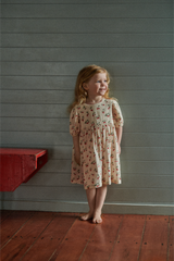 Young girl in a strawberry dress standing against a gray wall with a red shelf.