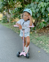 Child riding a pink scooter with a helmet on a path surrounded by greenery
