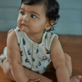 Baby sitting on a wooden floor wearing a sleeveless romper with sailboat pattern.