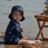 Baby in a navy swimsuit and hat sitting on a beach.