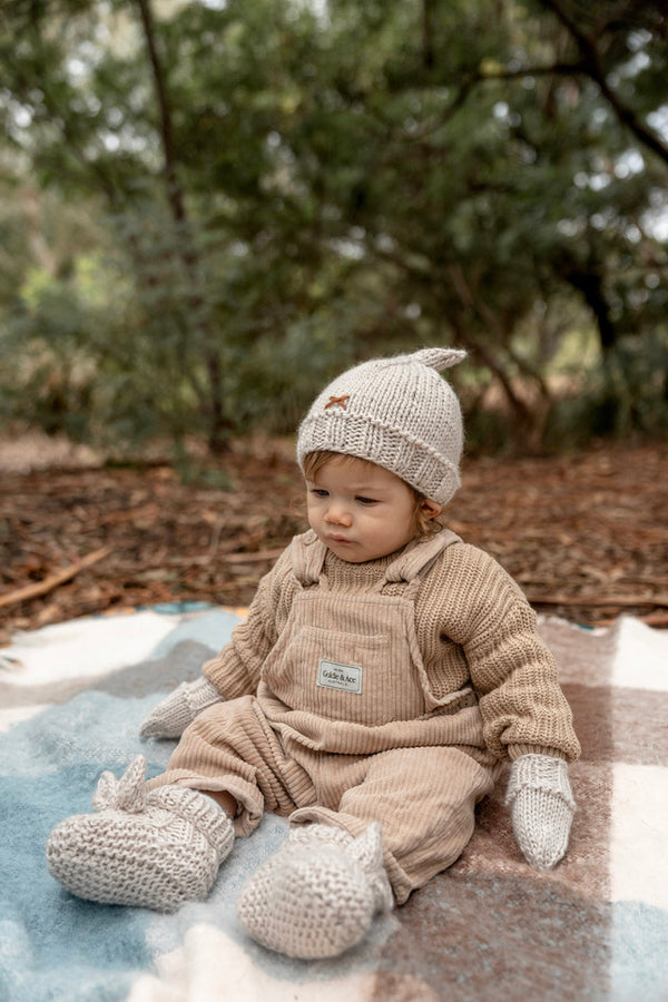 Baby sitting on rug wearing ACORN Cottontail Beanie Oatmeal and booties