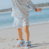 Child wearing a light blue checkered swim set with heart patterns and blue sandals on a sandy beach.
